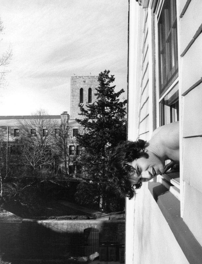 Black and white portrait of a young person leaning out of a window overlooking a building and trees on a bright day.