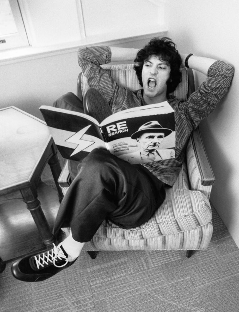 Adult male sitting in a striped armchair indoors reading a magazine with an image of a man wearing a hat, while yelling with his mouth open.