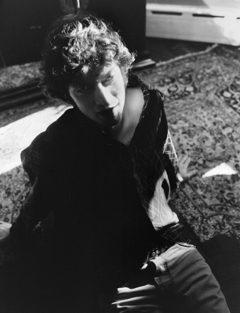 Monochrome portrait of a young adult male with curly hair, sitting on an patterned rug, looking slightly up with soft, natural window light.