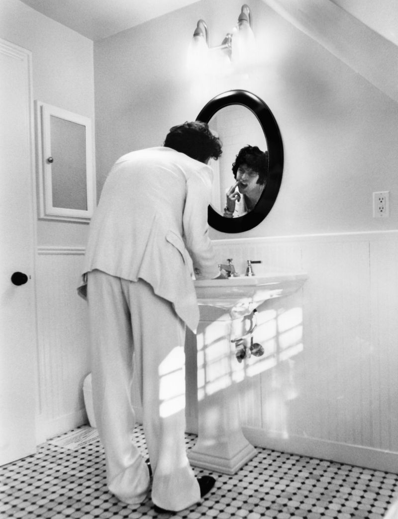 Black and white shot of person in white suit applying lipstick in bathroom mirror, vintage aesthetic with patterned tile floor.