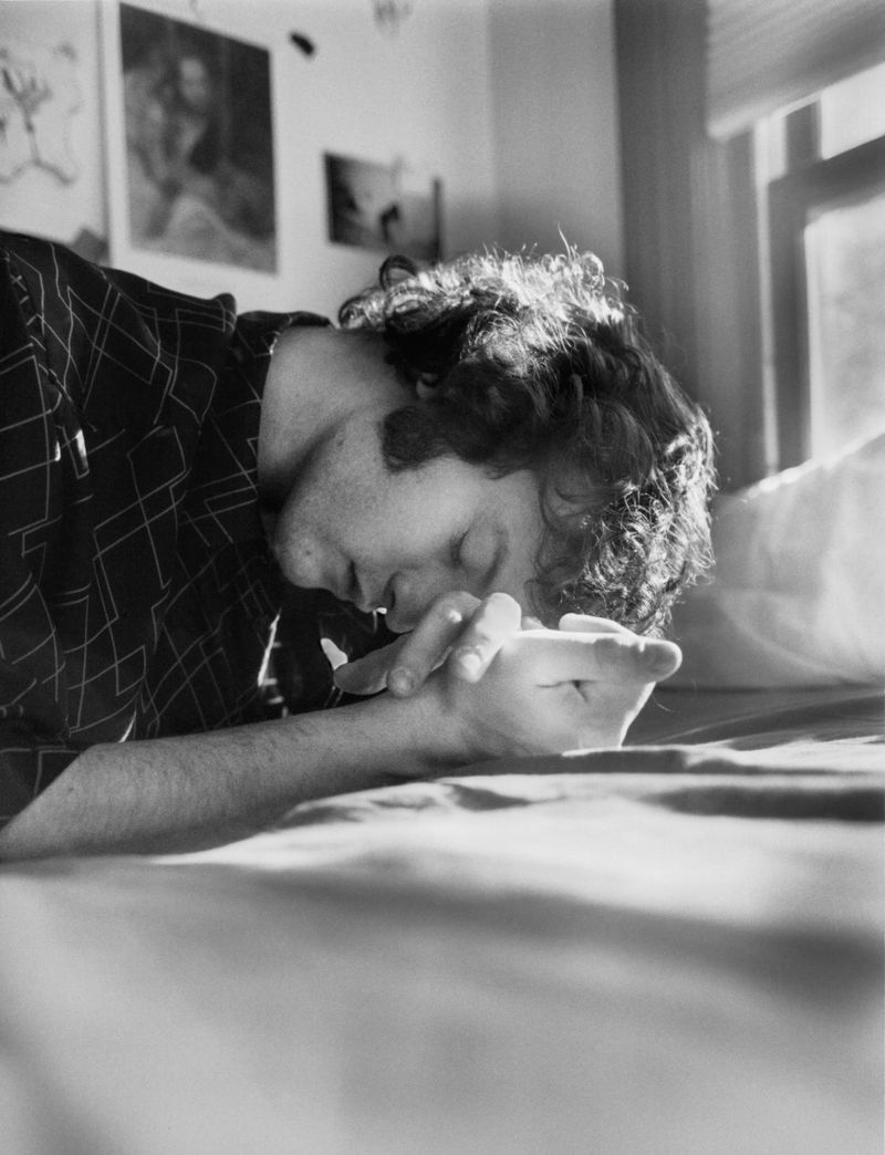 Monochrome portrait of a man lying on a bed with hands clasped, curly hair, eyes closed, wearing a patterned shirt, window light.