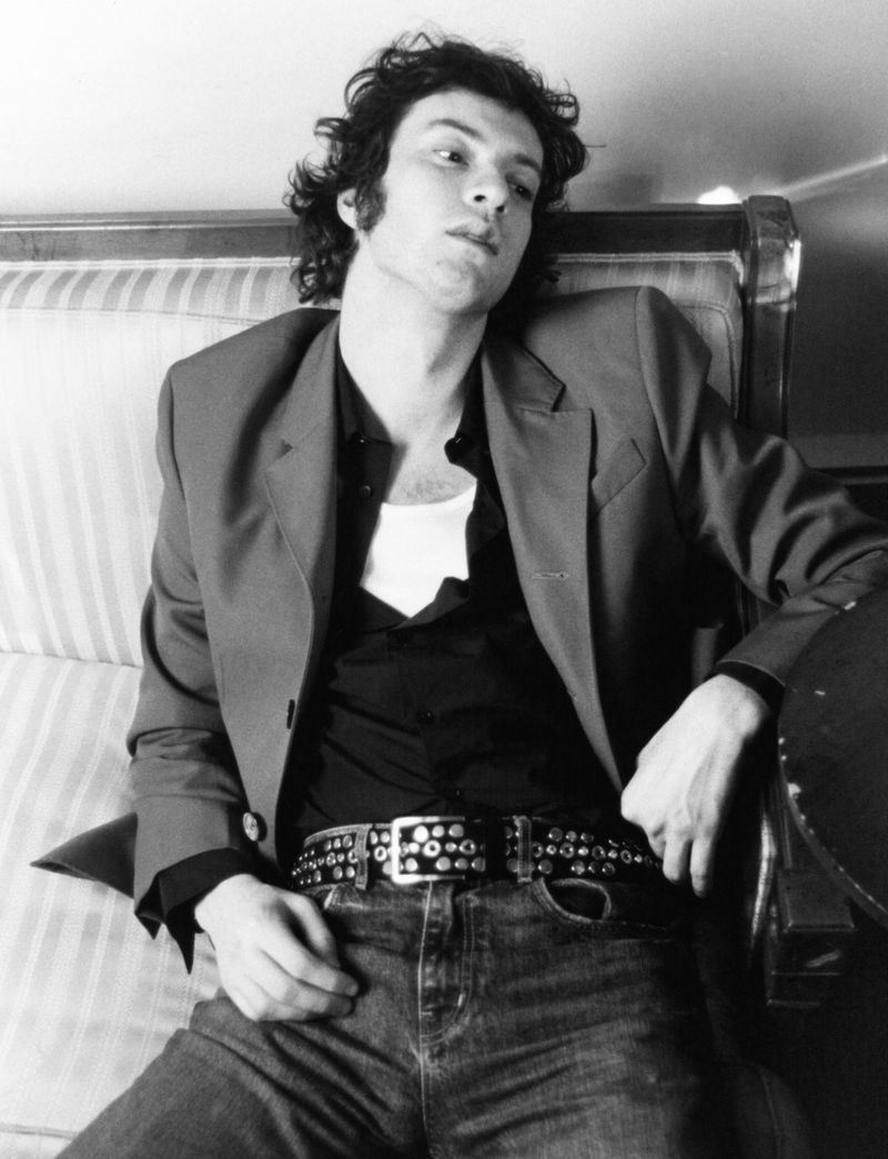 A man lounges on a striped cushion, wearing a blazer, shirt, jeans, and a studded belt, in a black and white portrait indoors.