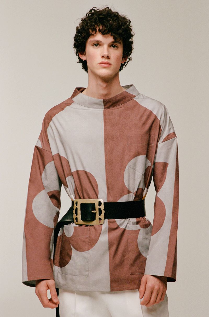 An adult male with curly dark hair poses in a photo studio wearing a color-blocked flower-patterned shirt and a black belt with a gold buckle.