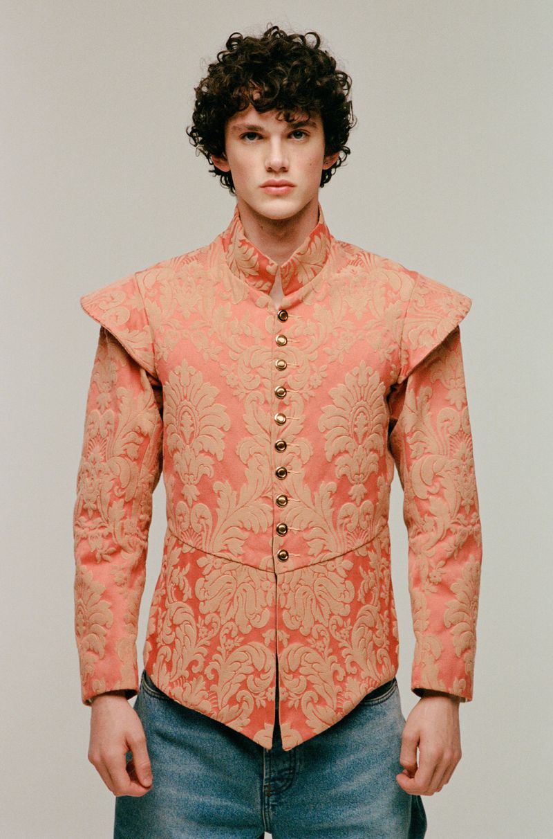 A young adult male with curly hair stands in a photo studio wearing a salmon and gold patterned top and light-wash jeans, posing for a portrait.