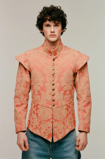 A young adult male with curly hair stands in a photo studio wearing a salmon and gold patterned top and light-wash jeans, posing for a portrait.