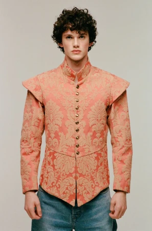 A young adult male with curly hair stands in a photo studio wearing a salmon and gold patterned top and light-wash jeans, posing for a portrait.
