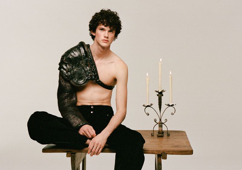 Young adult male with curly hair poses on a wooden table wearing partial armor and velvet pants, next to a candleholder with lit candles.
