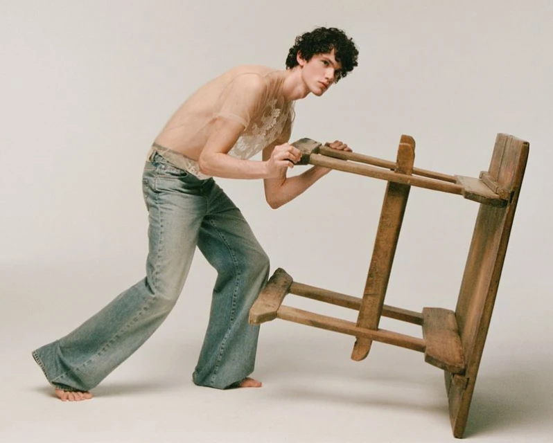 Young adult male model poses indoors wearing a translucent shirt and jeans, pushing an unfinished wooden easel against a white background.