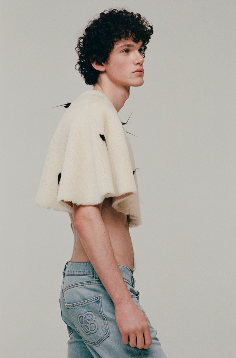 Young adult male model in denim jeans and ivory woolen cape posing indoors, studio shot. Fashion editorial, light background, portrait.