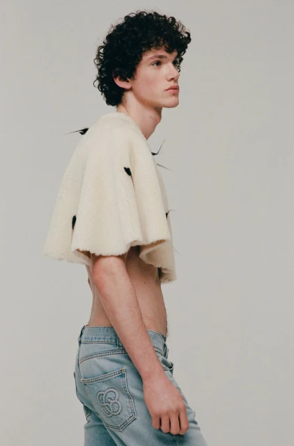 Young adult male model in denim jeans and ivory woolen cape posing indoors, studio shot. Fashion editorial, light background, portrait.