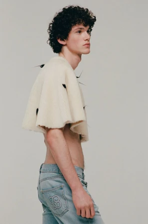 Young adult male model in denim jeans and ivory woolen cape posing indoors, studio shot. Fashion editorial, light background, portrait.