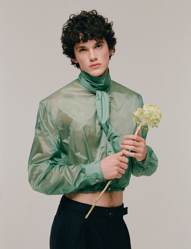 Young man with curly hair poses in sheer green shirt holding a white flower in a studio setting against a neutral background.