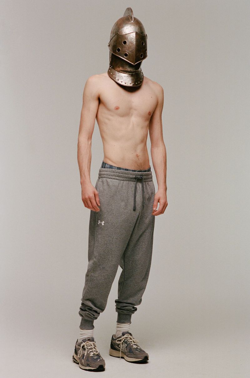 A shirtless man in gray sweatpants and sneakers stands in front of a plain backdrop, wearing a medieval knight's helmet.