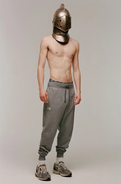 A shirtless man in gray sweatpants and sneakers stands in front of a plain backdrop, wearing a medieval knight's helmet.