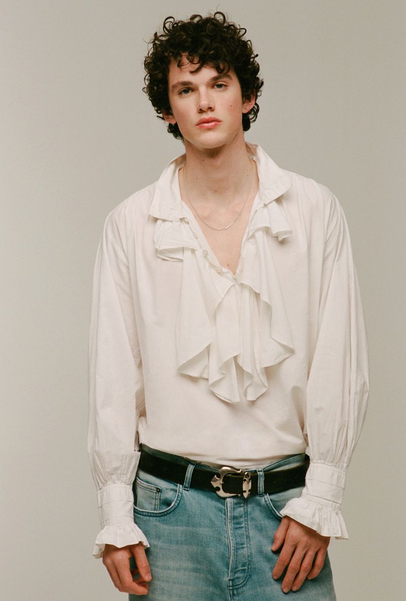 A young adult male poses indoors wearing a white ruffle shirt and blue jeans with a belt, against a neutral background in portrait orientation.