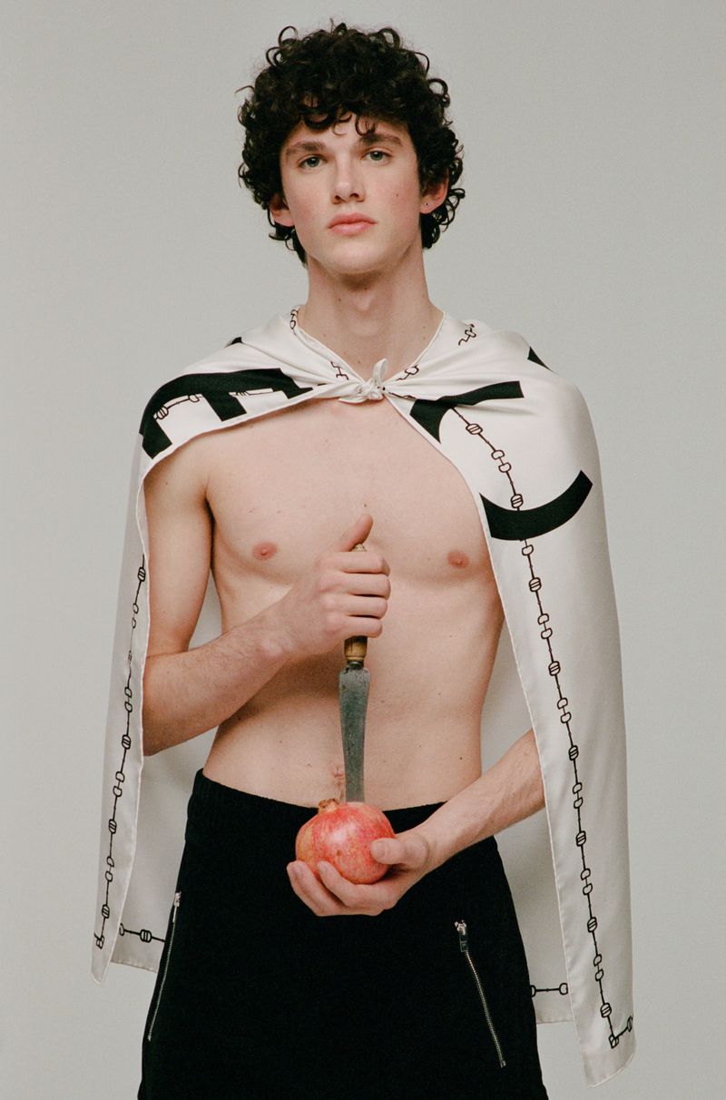 An adult male poses with a knife and pomegranate. He is shirtless, wearing a patterned cape, black pants, and facing the camera.