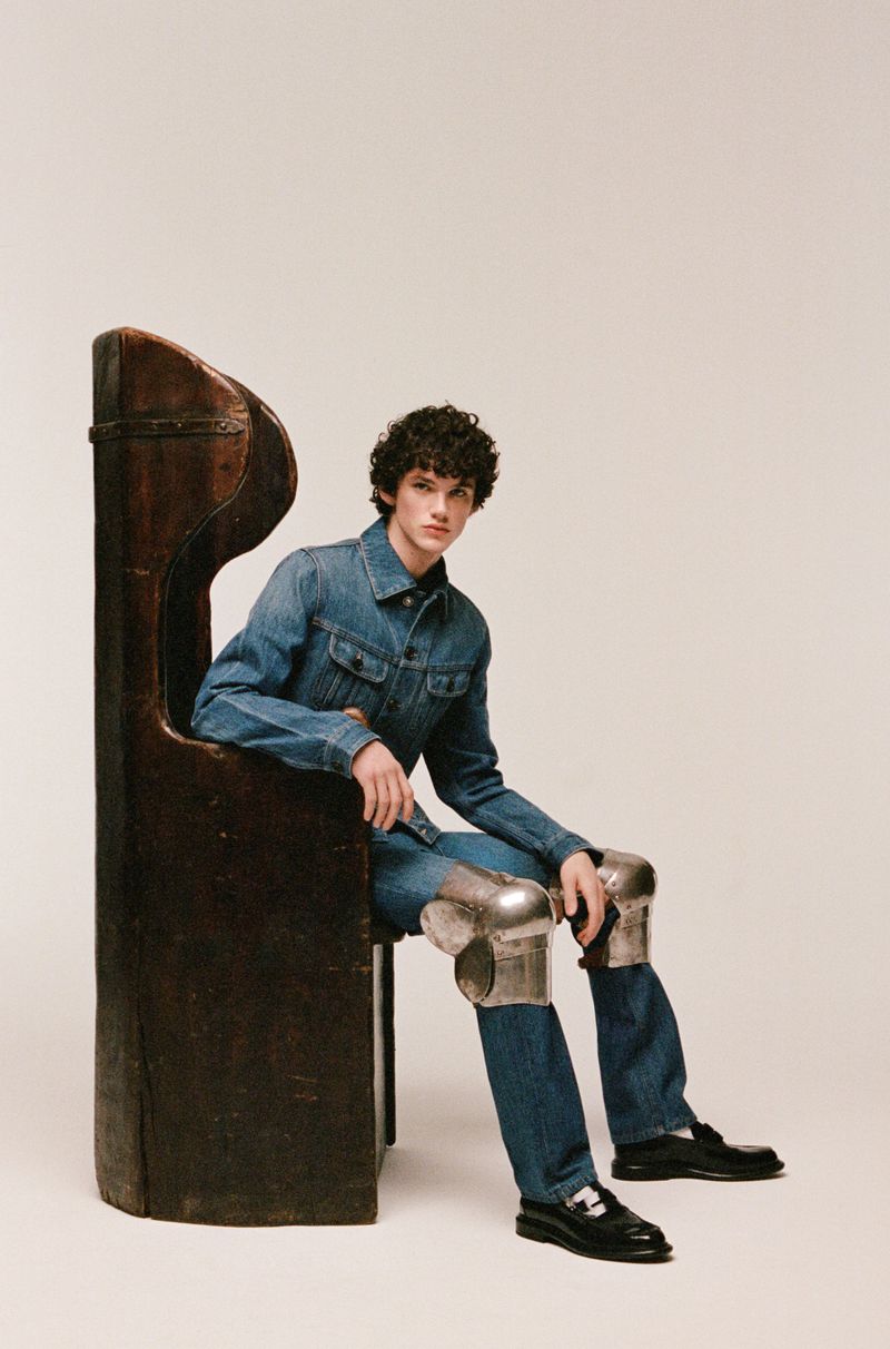 Man in denim jacket and jeans seated on a dark brown wooden chair with silvery knee pads, set against a neutral backdrop.