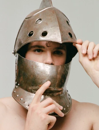 Man wearing antique helmet looks into the camera, holding the face plate in place. Indoor studio shot with neutral background.