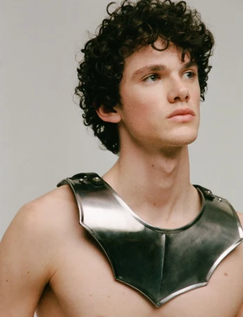 Young man with dark curly hair wearing a metallic shoulder armor piece against a grey background