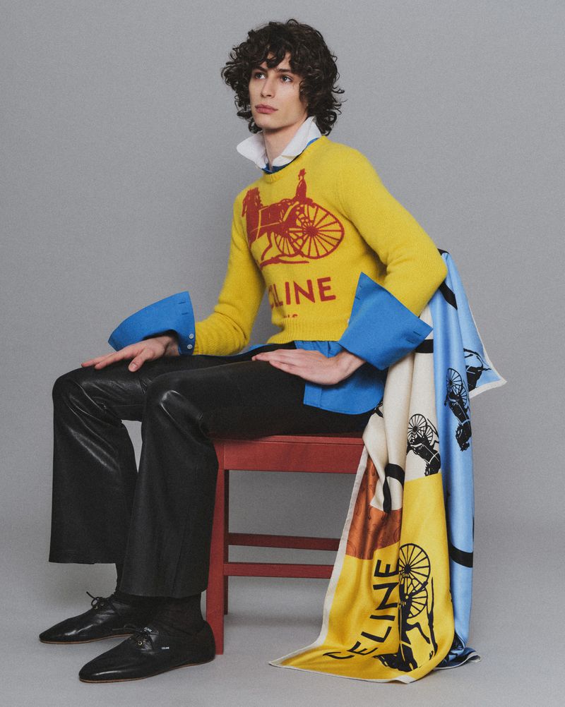 Fashionable adult male model with curly hair poses sitting on a red chair wearing a yellow sweater and black shiny pants in a studio.