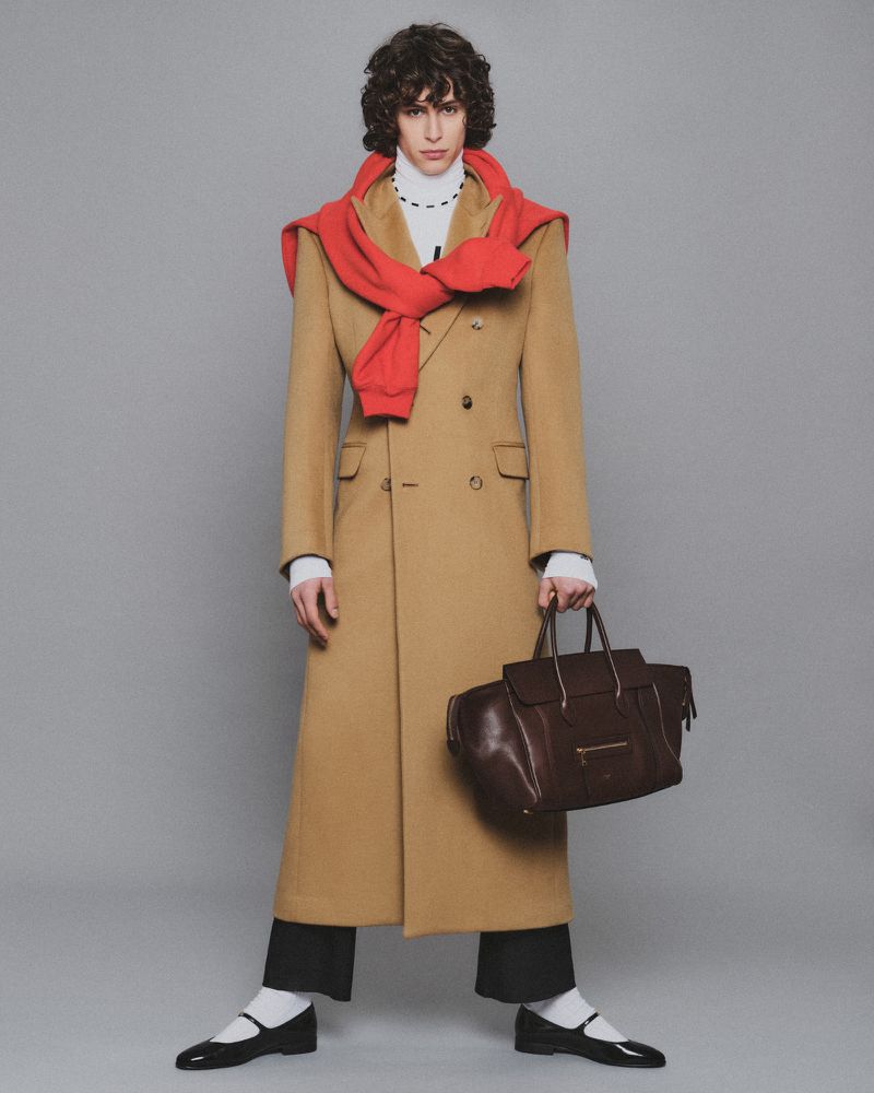 A slender man in a tan overcoat and black shoes stands against a gray background, holding a dark brown leather bag. Fashion shot.