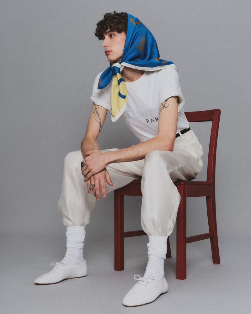 Young man with curly dark hair wearing a blue silk headscarf, white branded t-shirt, and white tapered trousers sitting on a red wooden chair.