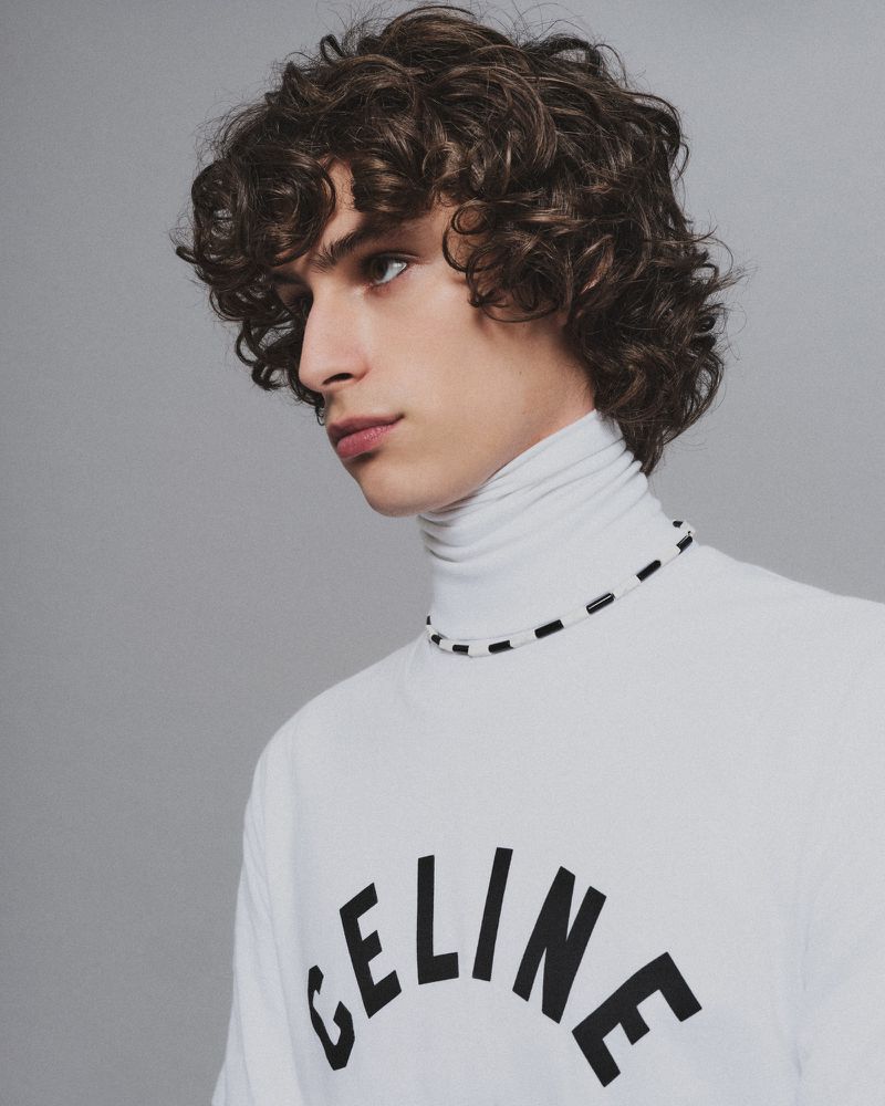 Portrait of a young adult male with curly hair, wearing a white turtleneck and necklace, against a neutral gray background.