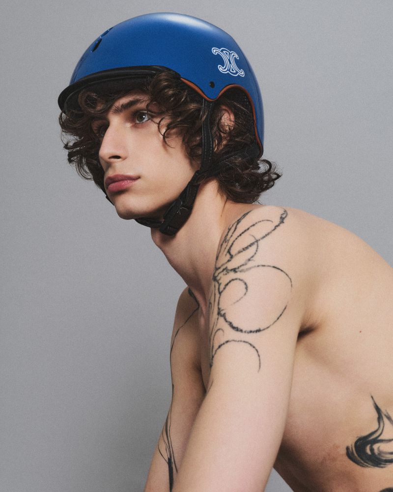 Studio portrait of a young adult male model with curly hair wearing a blue helmet with a logo and arm tattoos against a grey backdrop.