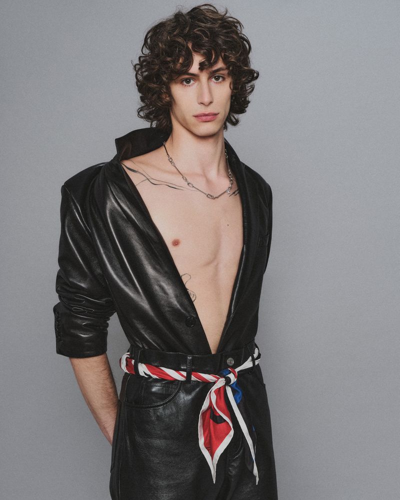 A young male model wearing an open black leather jacket with a tied red, white, and blue scarf belt against a plain grey studio backdrop