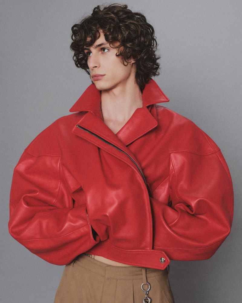 Adult male fashion model posing in a studio wearing a red leather jacket and beige pants against a grey backdrop. Fashion editorial.