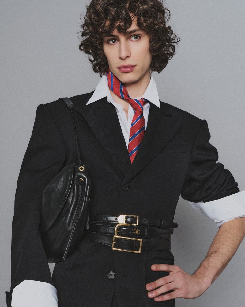 Young adult poses indoors wearing a black suit jacket, white collared shirt, striped tie, black belts, black handbag, and dark curly hair.