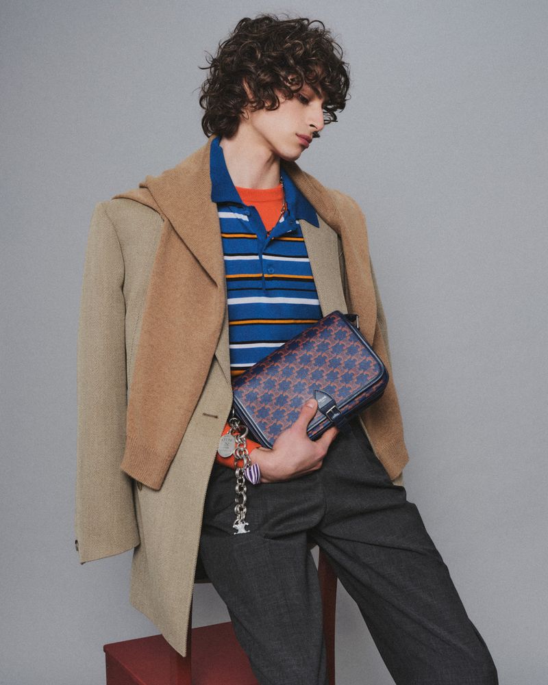 Adult male model poses indoors wearing a tan blazer, striped polo shirt, and grey pants, holding a patterned bag. Fashion editorial.