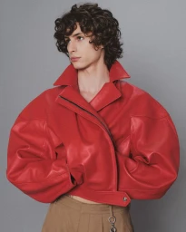 Young man with dark curly hair wearing a vibrant red leather jacket and beige trousers against a plain grey backdrop.