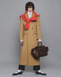 Model in a long tan coat with a red sweater draped over shoulders holding a dark handbag against a grey backdrop.
