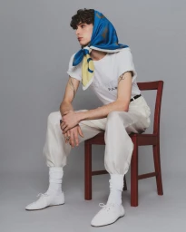 Young man wearing a blue silk scarf, white t-shirt, and white trousers seated on a red wooden chair against a grey studio background