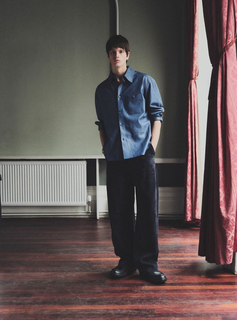 A stylish adult male stands indoors with a blue collared shirt and dark pants, with a radiator and red drapes in the background.