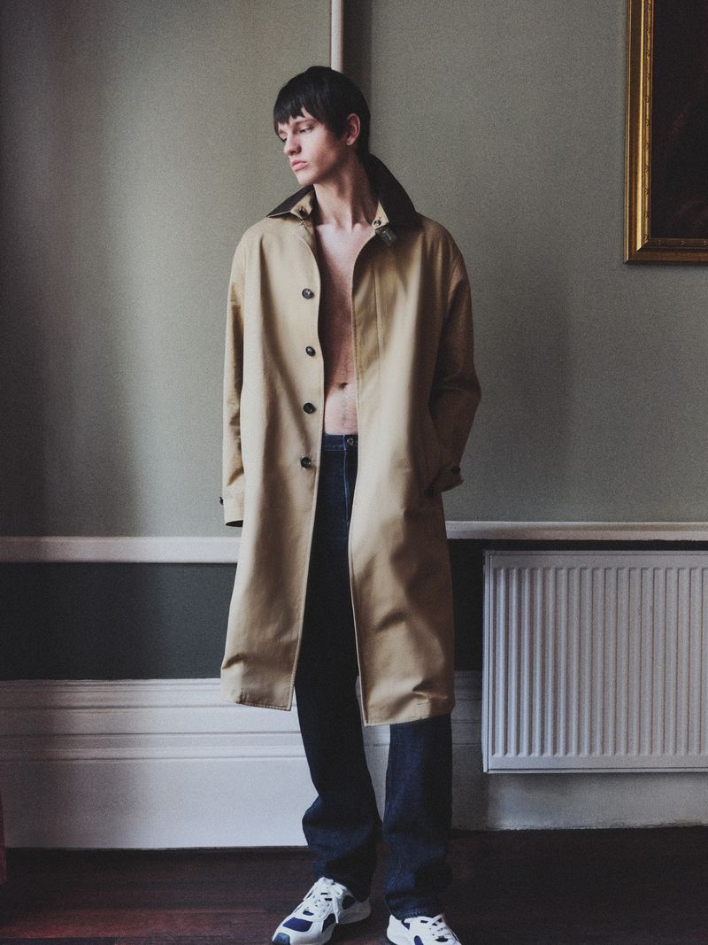 A slender adult male model stands in a room with a radiator, wearing a long beige coat, jeans, and sneakers, posing in soft window light.