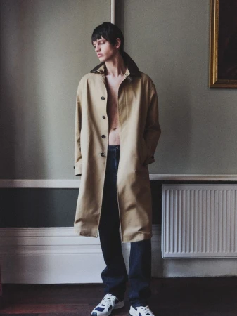 A slender adult male model stands in a room with a radiator, wearing a long beige coat, jeans, and sneakers, posing in soft window light.