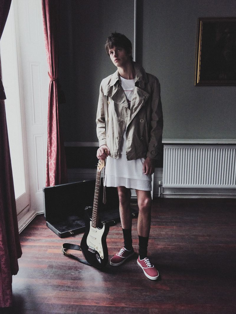 Stylish young man stands indoors with an electric guitar, wearing a beige jacket, white dress, red sneakers; soft light from the left.