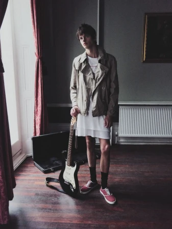 Stylish young man stands indoors with an electric guitar, wearing a beige jacket, white dress, red sneakers; soft light from the left.