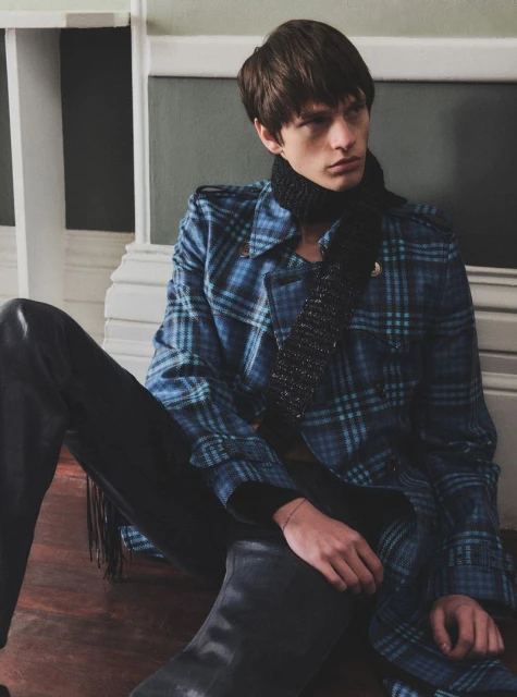 Young adult man in a blue plaid coat and black scarf sitting indoors near a wall, fashionably posed with dark pants and boots.