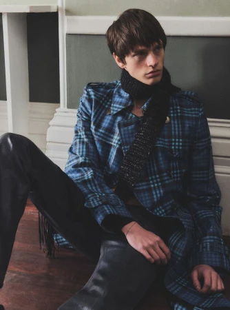 Young adult man in a blue plaid coat and black scarf sitting indoors near a wall, fashionably posed with dark pants and boots.