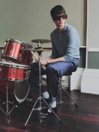 Male musician wearing sunglasses seated with drumsticks in hand, with a red drum set in a room with neutral background and lighting.
