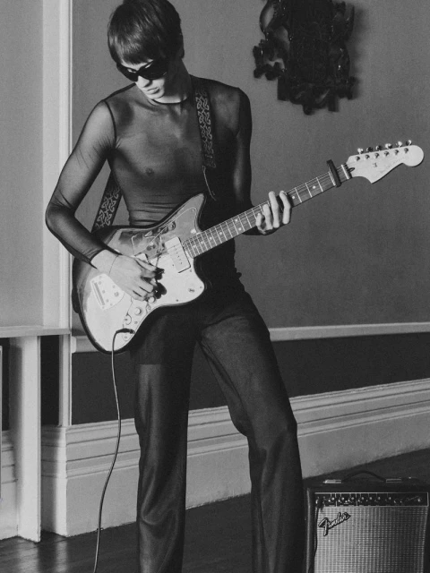 A musician in a sheer top and sunglasses plays an electric guitar in a room with neutral tones, with a Fender amplifier in view.