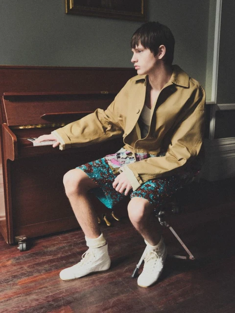 Young adult male seated at a brown upright piano, indoors, wearing a tan jacket, patterned shorts, white socks, and white high-top sneakers.