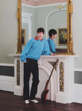 A young adult male in a blue sweatshirt leans against a white fireplace with a mirror, with an electric guitar propped beside him indoors.