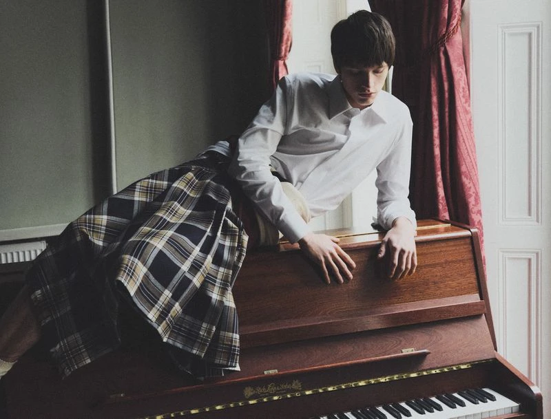 A young man in a white shirt and plaid skirt leans on a wooden piano in a room with natural light and red curtains.