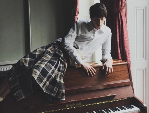 A young man in a white shirt and plaid skirt leans on a wooden piano in a room with natural light and red curtains.