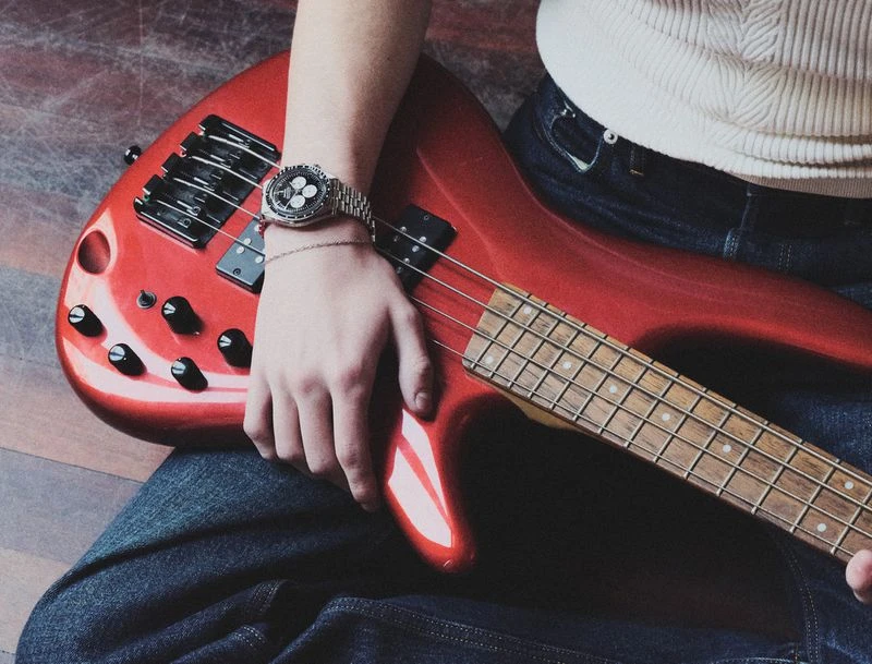 Close-up of adult wearing a watch and blue jeans, resting hand on a red electric bass guitar against a wood-patterned surface.