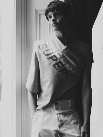 Monochrome fashion shot of a young adult man indoors wearing a branded t-shirt and light-colored trousers, near a window with dark shadows.
